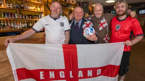 City of Wolverhampton Council Four men are standing together, with the man on each end of the line holding an England flag and wearing an England top. They are in a bar setting and the man second from the left is wearing a jacket, shirt and tie.