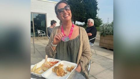 Liza Raby, 54, holding her medal and smiling while eating fish and chips. 