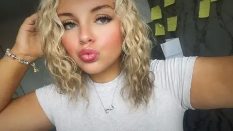 Megan Swann is looking at the camera. She has wavy blonde hair and is wearing a pale T-shirt