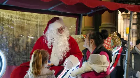 City of Wolverhampton Council Father Christmas speaks to a woman with dark hair who is carrying a baby and accompanied by another young child
