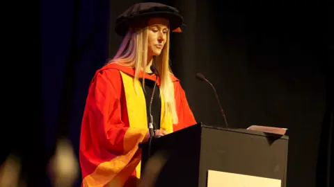 University of Wolverhampton Hampton is standing side on to the camera at a mainly black lectern that includes a black microphone. She is wearing a red and yellow gown and looking down, in front of a black background.