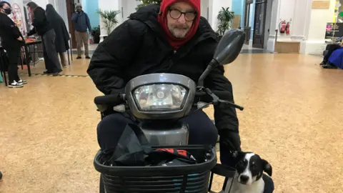 Malcolm McCormick visiting 2 Royal Avenue with his pup Chelsea.  Malcolm is using a mobility scooter and is wearing a black coat, glasses and a red snood.  The pup has black, white and tan markings. 