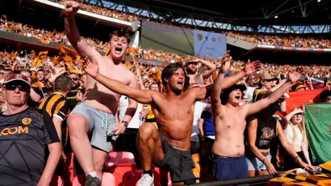 PA Three shirtless young men with their arms spread wide in celebration stand at the front of a crown of Hull City fans
