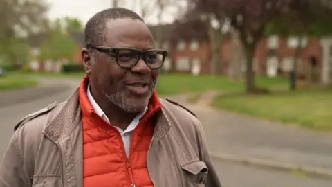 A man in an orange zipped sweater and a beige jacket stands on the pavement on a residential street 