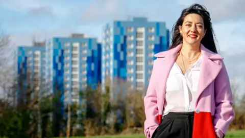 Sarah stands outdoors in a park with trees at the edges, behind which are the blue tower blocks of Marsh Farm. She wears a white blouse under a pink and red open coat, and has long dark hair.