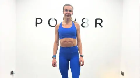 POW8R Holly wears a Colbalt blue gym set which includes gym legging and sports bra. She has her hair in a pony and is stood in her home gym