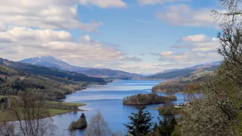 Ryan Gearing A view along a lake with hills and trees either side under a cloudy blue side.