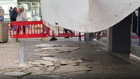 A large white stretch of plasterboard has fallen from a row of shops, and there is debris all over the ground.