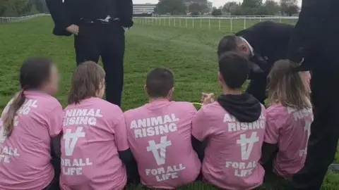 A group of five people wearing matching pink shirts that read “ANIMAL RISING – FOR ALL LIFE” sit on a grassy racecourse. They face several individuals in dark suits who appear to be engaging with them.
