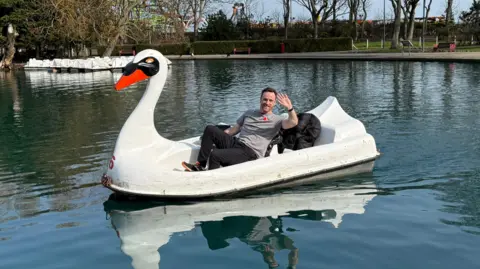Matt Bailey has short brown hair and is wearing a grey t-shirt with black trousers waves while sitting aboard a giant swan-shaped pedelo on a lake. The sky is blue and the water ripples slightly in the breeze.