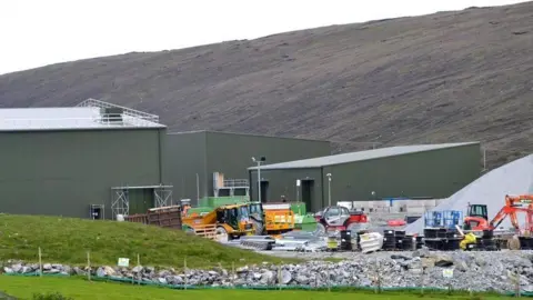 Three corrugated metal-walled buildings dominate the site. There is a yard with construction vehicles, including a tipper truck and excavators.  