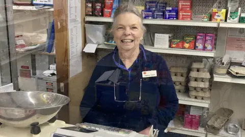 An older woman with white hair and a dark blue jumper smiles at the camera from behind a till in a village shop