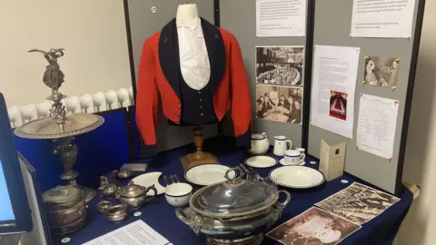 A red and black soldier's jacket which would have been worn to a mess hall banquet. Around it are white and blue china plates and cups, with a large silver serving pot.