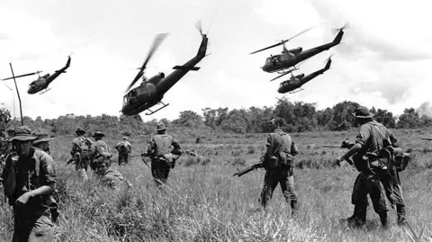 Getty Images Australian troops carrying guns walk through long grass. Four helicopters fly low over the terrain.