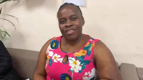 Norah Kiyika, who is wearing a pink dress with colourful flowers, is sitting on a couch and smiling at the camera. 