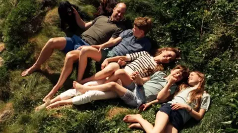 The Prince of Wales lies in the grass in a khaki tshirt and blue shorts, his hand is resting on his black spaniel dog, next to him is Prince George in a blue tshirt and green shorts, the Princess of Wales is smiling next to them in a white and navy stripped tshirt and white trousers, she holds the hand of Prince louis who wears a green shirt and chequered blue shorts, Princess charlottes is wearing black shorts and a pale green tshirt.