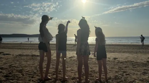 Four young girls are pointing up at the sky while standing on a sandy beach