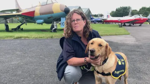 A woman in glasses, wearing a dark coloured top and light coloured trousers is looking at the camera and kneeling down next to a golden coloured dog.
The dog is wearing a vest that says Guide Dogs. In the background there is some grass with several planes on display at the museum.