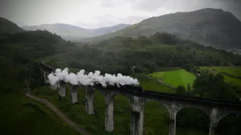 Colin Eaton White plumes of smoke rise from a train on a train track which is elevated by stone pillars. There are green fields and mountains all around it.