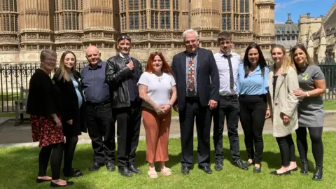 Ten people in a group picture taken on grass outside the Houses of Parliament in bright sunshine. A man with short grey hair and a navy blue suit, blue and white checked shirt and colourful tie stands in the centre next to a woman with shoulder length red hair, wearing a white t-shirt and rust coloured wide trousers and white flat sandals.