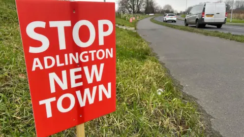 A sign next to a busy road which says stop Adlington new town