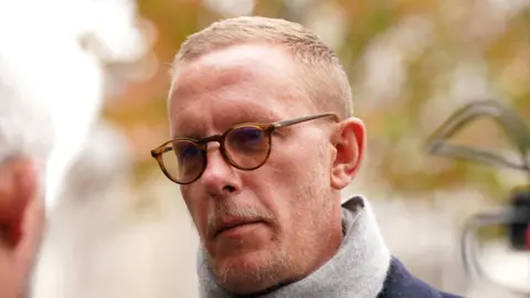 PA Media Laurence Fox has short blond hair and stubble. He is wearing round-framed brown glasses and a grey scarf over a navy blue coat.