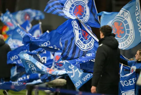 Getty Images Rangers flags being waved by fans with one man in a dark jacket standing in front of them