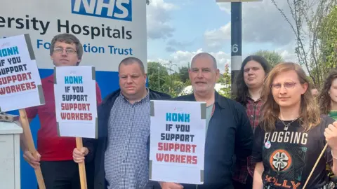 A group of six people from Unite the Union outside Addenbrooke's Hospital in Cambridge. They are holding placards saying honk if you support support workers. A sign saying NHS can be glimpsed on the left behind them. 