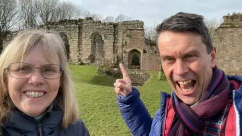 Paul Silvester Paul and his wife are grinning at the camera, with a ruin in the background