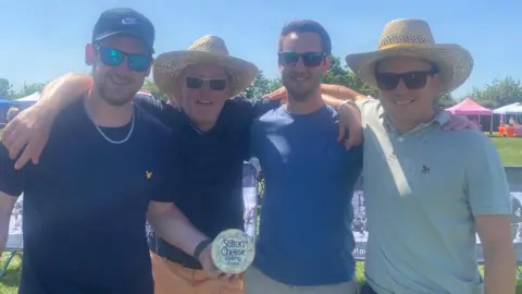 A group of four men stand together with their arms around each other and smile at the camera. One on the end holds a small wheel of Stilton cheese. Two of the men wear large hats and another wears a cap. They are all wearing sunglasses and T-shirts.
