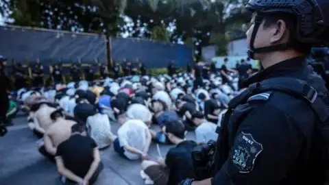 Petugas Polisi Kokang berdiri mengelilingi puluhan pria yang duduk di tanah, dengan tangan terikat di belakang punggung.