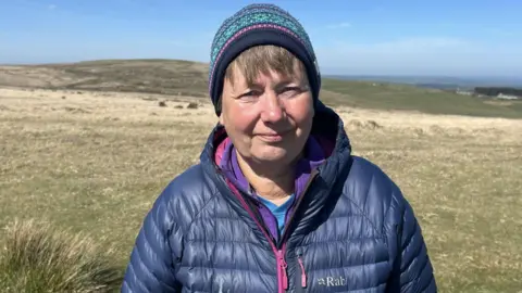 A woman, wearing a colourful woolly hat, is smiling at the camera. She is wearing a dark blue puffer jacket with a pink zip, and she is standing on moorland.