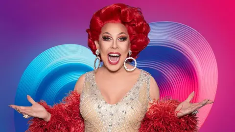 Am image of La Voix in a sparkly dress and red feather boa and bright red hair, in front of the Eurovision colourful heart logo