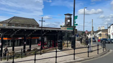 Carmelo Garcia A street view with a shelter, pavement and clock tower. Someone is stood beneath the shelter and there is another person sat on a bench on the pavement. There are a couple cars on the road. The sky is blue with some clouds. 