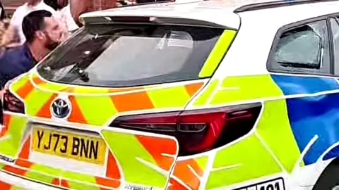 West Yorkshire Police A man standing by the rear left window of a police car, which has its window on the other side smashed. His body is facing to his left.