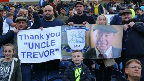 Getty Images Supporters of Preston North End pay tribute to former owner Trevor Hemmings during the Sky Bet Championship match between Preston North End and Derby County at Deepdale Stadium on 16 October 2021. They hold a large banner, featuring a smiling photograph of Mr Hemmings wearing a flat cap. The message "Thank you 'Uncle Trevor' is printed next to the club badge.
