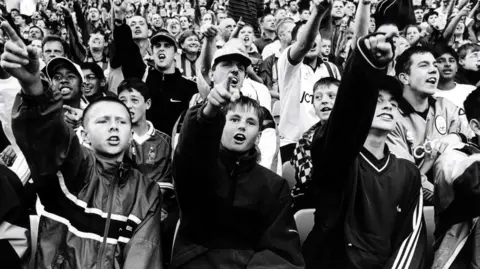 Ian Beesley A black-and-white photo of a football crowd with three teenage boys in tracksuits at the front pointing into the air.
