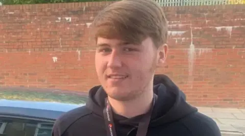 Ellis Cox stands in a black hoody in front of a car and wall 