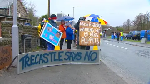 Protesters in Trecastle holding umbrellas and placards saying "Trecastle says no" next to the roadside