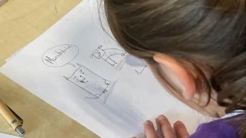 A girl in a purple school jumper draws a cartoon picture of a dog and a cat.