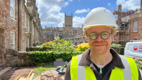 Nik Kitchridge is standing outside a house on Vicars' Close. He is wearing a white hard hat and a hi-vis jacket. Wells Cathedral is in the background. Scaffolding is on a row of houses.
