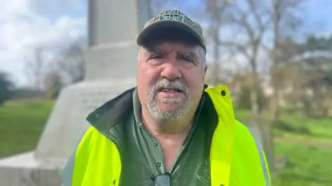 Steve Davies is dressed in a high vis jacket and cap, looking to camera. He is standing in front of the memorial in the cemetery.