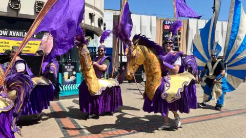 Emma Baugh/BBC Men wearing purple shoulder pads and hats with purple feathers. They are in outfits that make them look like they are riding atop golden horses. 
