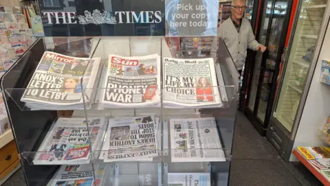 Gordon Bird A large wrack of newspapers on display in a newsagents. An elderly man in a grey cardigan stands beside them