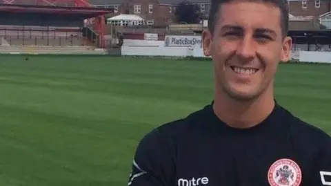 Accrington Stanley FC A man in a football kit for Accrington Stanley standing on a pitch, smiling. He has brown hair. 