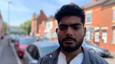 Farhan Rafik is pictured wearing a white shirt and grey light coat. He has a beard and cars and houses are blurred in the background