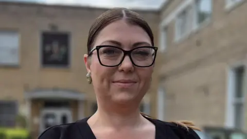 Stuart Woodward/BBC Danielle Belton has her brown hair in a ponytail and is wearing glasses with black rims. Behind her is a cream building.