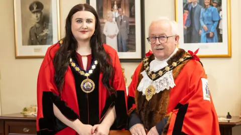 Mid and East Antrim Borough Council Bethany Carson-Ferris and Thomas Gordon are wearing official red robes and gold chains of office. A picture of Queen Elizabeth and Prince Philip, and a picture of Prince William and Princess Kate are behind them. 