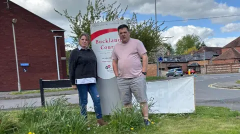A man with brown hair and wearing a pale pink T-shirt and grey shorts is pictured standing next to his wife on a grass verge by the side of a road. The woman has blonde hair and is wearing a dark grey jumper and dark blue jeans. Both are standing in front of the 'Buckland Court' care home sign on the road side with houses and parked cars in the background.