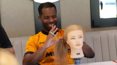 Wolves Foundation A man in a Wolves top battles with a hair bobble using a plastic hair model on a table in front of him.